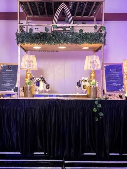 A stylish and modern bar setup for a cocktail party. The design features geometric accents, lush greenery, and elegant lamps, creating a sophisticated spot for guests to grab a drink.