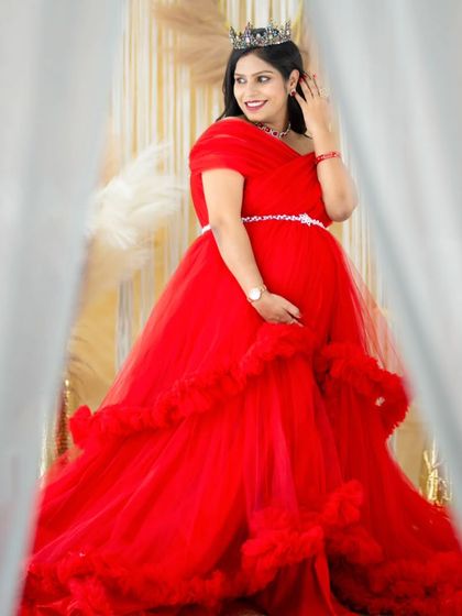 A candid moment of joy. The mom-to-be, wearing a vibrant red gown, looks off-camera with a beautiful smile, framed by the soft curtains of our studio.