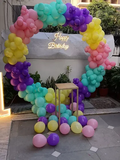 This outdoor first birthday setup uses a circular balloon arch in bright, playful colors. The arrangement includes a 'Happy Birthday' neon sign and a large marquee number '1', designed to stand out in any backyard or garden party.