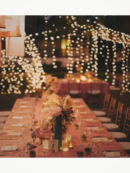 A magical evening setting with long tables adorned with flowers and candles, set under a canopy of warm, cascading string lights. This creates an intimate and enchanting atmosphere for any celebration.