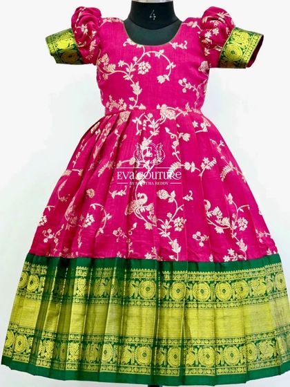 A vibrant pink frock made from floral Banarasi fabric. The puff sleeves and rich green and gold border make it a festive choice.