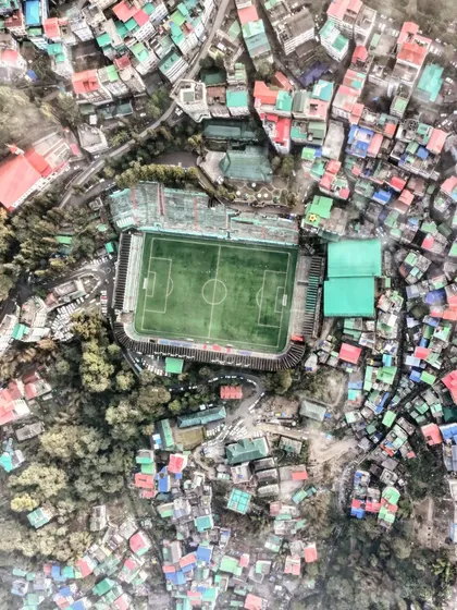 A top-down view of a football stadium in Gangtok, Sikkim, surrounded by a dense, colorful cityscape. This shot highlights the challenge of urban planning in a hilly region.