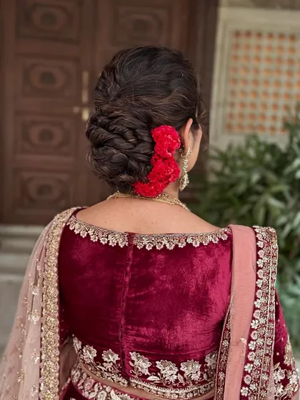 A detailed view of a royal yet modern messy classic updo. This intricate hairstyle, adorned with fresh red flowers, was designed to complement the client's velvet lehenga and add a touch of timeless elegance to her reception look.