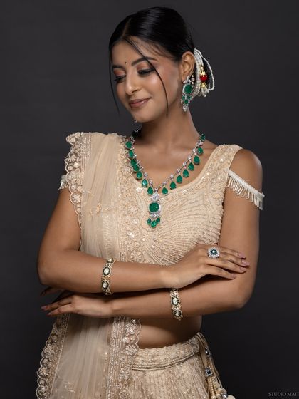 A model in a cream lehenga, showcasing a beautiful emerald jewellery set.