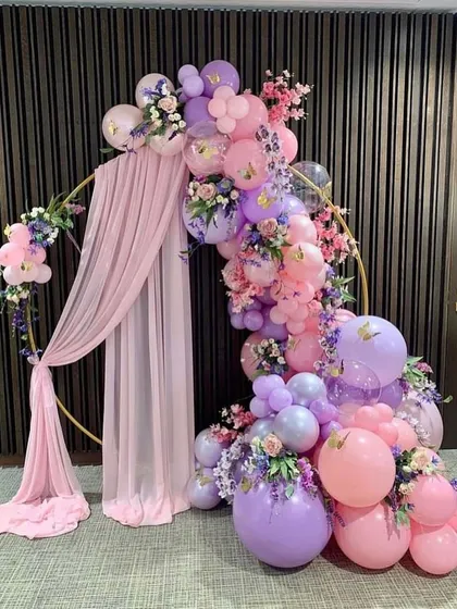 An elegant circular hoop decorated with pink and purple balloons, artificial flowers, and draped fabric, creating a sophisticated photo backdrop.