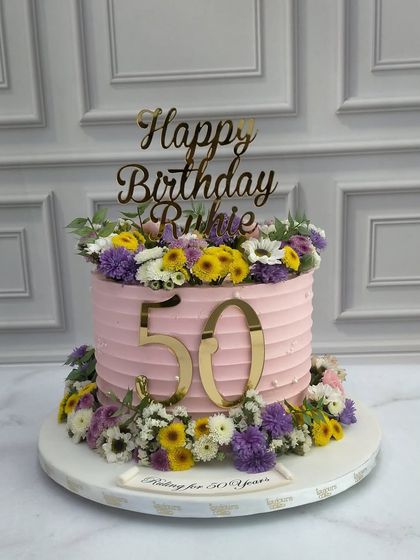 Celebrating a 50th birthday with a burst of color. This cake features a pink ribbed buttercream finish and is adorned with a crown and base of fresh, vibrant wildflowers in shades of purple, yellow, and white.