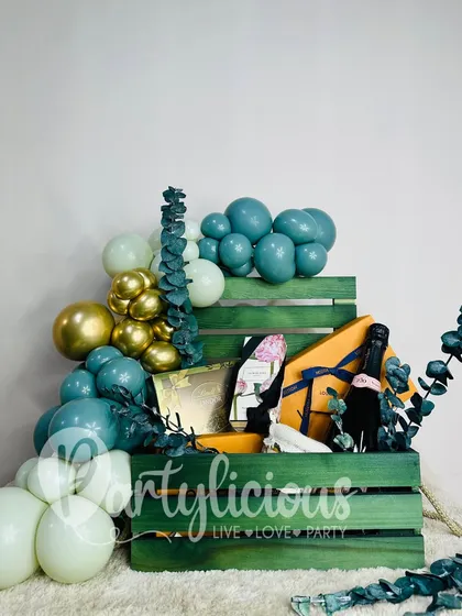 A luxurious gift hamper in a teal-painted wooden crate. The gifts are nestled amongst a cascade of teal and gold balloons, creating a high-end, beautifully styled present.