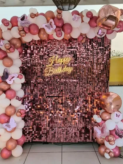 A terrace party with a rose gold sequin wall. The setup features a "Happy Birthday" neon sign and a balloon arch in shades of pink and white with butterfly accents.