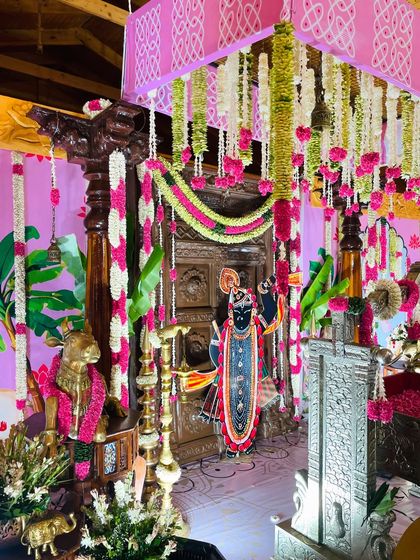 A full view of the vibrant and devotional naming ceremony setup. The entire space is filled with color, from the pink canopy and floral garlands to the traditional artwork and brass lamps.