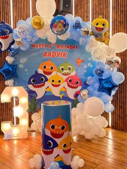 An underwater adventure with a Baby Shark theme for a 3rd birthday. This setup includes a custom backdrop, character foil balloons, a light-up number '3', and a blue and white balloon frame.
