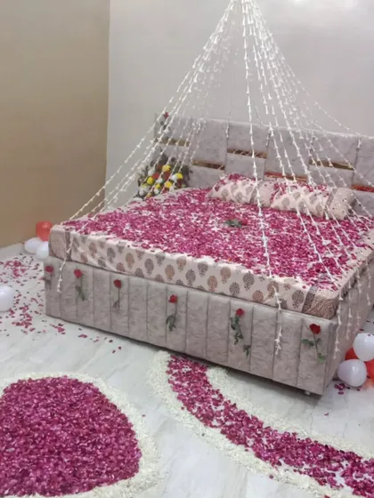 A simple yet beautiful first night room decoration. The bed is covered in pink rose petals, with a delicate canopy of light strings above. A heart and path are also created on the floor using petals.