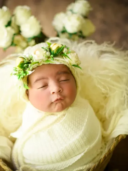 A close-up on a sleeping baby girl wearing a beautiful floral crown, wrapped in a cream-colored swaddle.