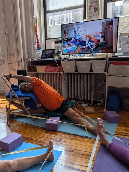 A hybrid class in session, with me demonstrating Parivrtta Parsvakonasana (Revolved Side Angle Pose) using a chair. The screen shows the online view for remote students.