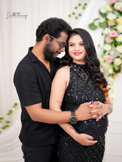 An intimate couple portrait against a floral backdrop. The husband embraces his wife from behind, both looking down at the baby bump with love and anticipation.
