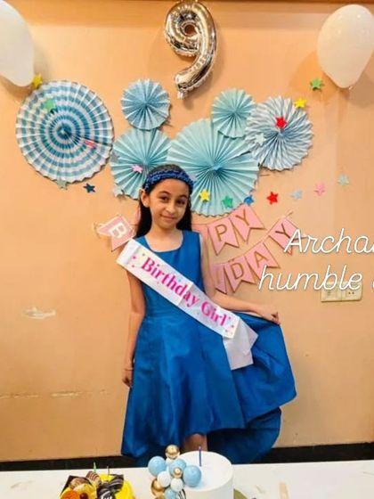 The birthday girl looking beautiful in her blue dress, standing by her balloon-themed cake.