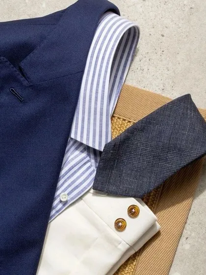 A flat lay of a classic menswear combination: a navy blazer, striped shirt, white trousers, and a textured tie. These are the building blocks of timeless style.