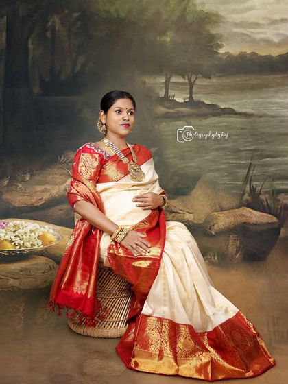 A peaceful studio portrait of a mother-to-be in a traditional red and white saree, seated against a painted backdrop.