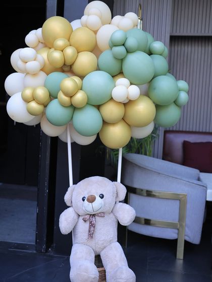 A close-up of a teddy bear hot air balloon decoration. The bear is lifted by a bundle of sage green, gold, and cream balloons, a popular element in my baby shower designs.
