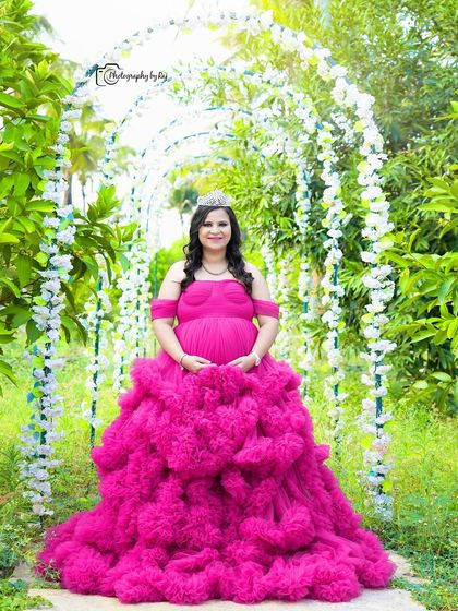 A "Barbie" themed portrait of a mother-to-be in a vibrant pink ruffled gown and crown, framed by a floral archway.