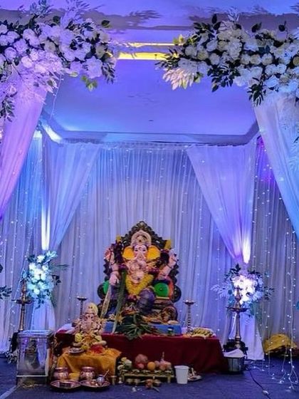 A grand and dramatic Ganpati setup with white floral arrangements and flowing white curtains. The cool blue uplighting gives the entire space a majestic and ethereal feel, perfect for a large hall.