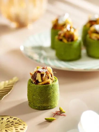 A macro shot of a modern, pistachio-based mithai from the Marriott Diwali collection. The soft lighting and shallow depth of field draw attention to the intricate textures and silver leaf garnish of this gourmet sweet.