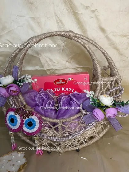 This purple-themed Diwali hamper is all about spreading joy. The jute basket is filled with a box of Kaju Katli and other surprises, decorated with floral accents and evil eye hangings.