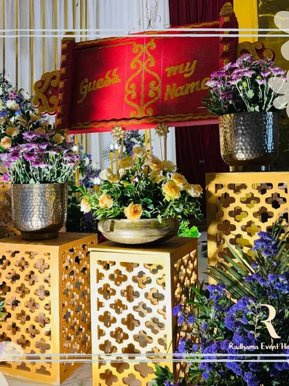 A close-up of the "Guess My Name" scroll, a fun and engaging element for a naming ceremony. The setup is decorated with intricate jaali pedestals and pots of fresh yellow roses and purple chrysanthemums.