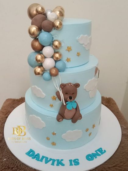 A sweet and classic choice for a first birthday, this three-tiered cake features a teddy bear theme. The design includes a fondant bear holding balloons, set against a sky-blue background with clouds and stars.