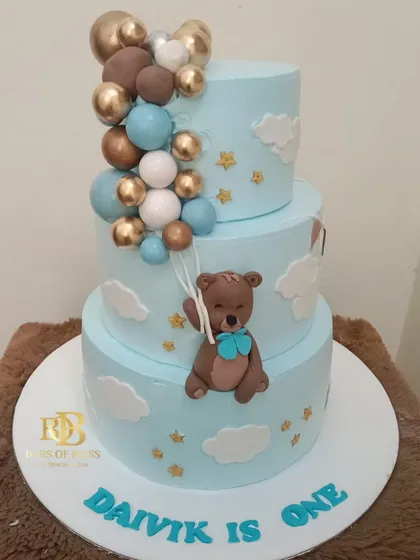 A sweet and classic choice for a first birthday, this three-tiered cake features a teddy bear theme. The design includes a fondant bear holding balloons, set against a sky-blue background with clouds and stars.