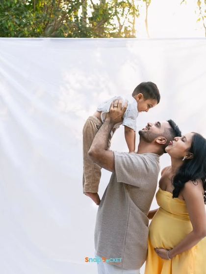 A father lifts his son high in the air while the mother looks on lovingly. This outdoor maternity photo is full of life and happiness.