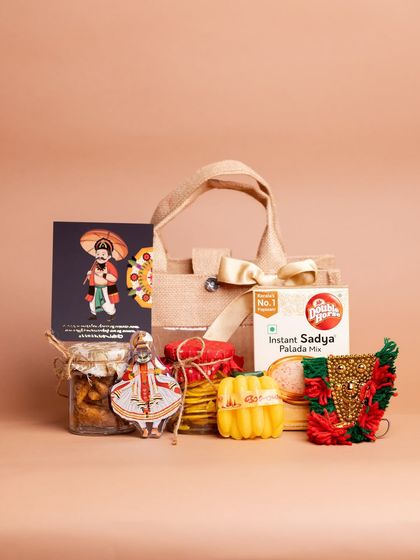 The full set of items from the 'Kaithola' hamper, displayed with the jute bag. It includes snacks, a payasam mix, and small cultural trinkets like a Kathakali magnet and a mini Nettipattom.