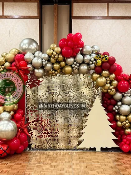 A festive Christmas-themed celebration with a gold shimmer wall and a balloon arch in red, gold, and silver. The Christmas tree cutout adds a perfect seasonal touch.