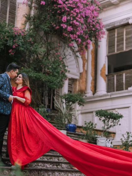The vibrant red of the gown stands out beautifully against the rustic charm of the building and the soft purple flowers. This is a perfect example of a romantic, timeless pre-wedding photo.
