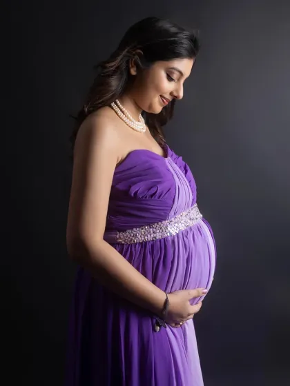 A profile shot capturing the beautiful curve of the baby bump. The mom-to-be wears a strapless purple gown with an embellished waistline, her gentle smile reflecting a moment of quiet joy.