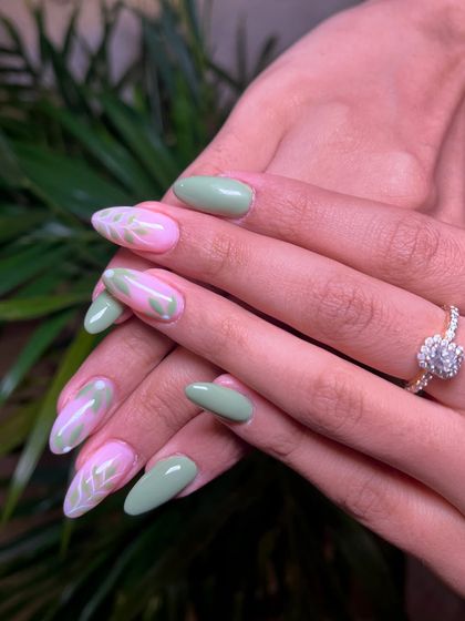 When elegance meets nature. This beautiful almond-shaped set combines solid pastel green nails with accent nails featuring a pink base and delicate white leaf patterns.