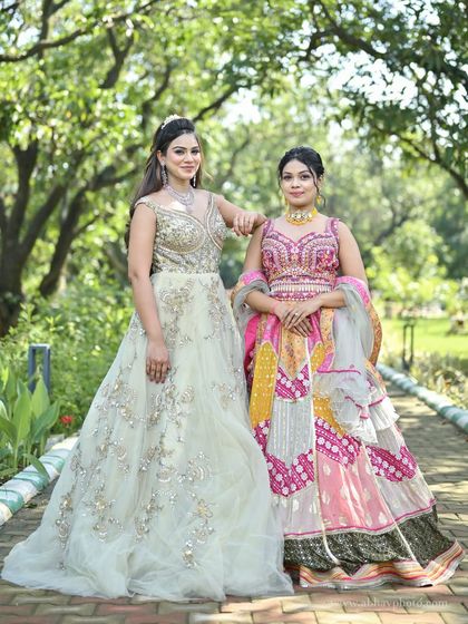 A friendly pose showing two distinct styles. The off-white gown with a diamond necklace contrasts beautifully with the bright, multi-colored lehenga, perfect for different wedding events.