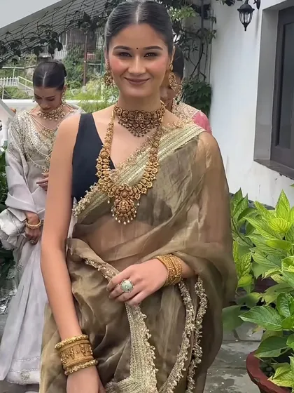 A model wearing a trending olive green organza saree, paired with a black blouse and heavy traditional gold jewellery for a statement look.