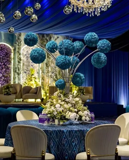 A dining table setup with a creative centerpiece of blue floral spheres. The deep blue tablecloth and surrounding drapes create a rich, monochromatic look.