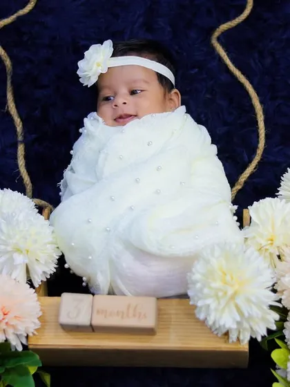 This newborn girl is wrapped in white and resting on a swing decorated with soft white and pink flowers, creating an angelic and dreamy portrait.