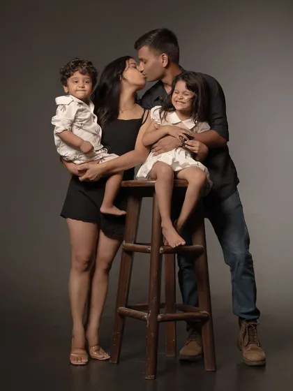 Against a classic dark studio background, a family of four comes together in a beautiful, candid embrace. The father's gentle kiss and the children's natural poses show that a family's love is a story told in small, authentic gestures.