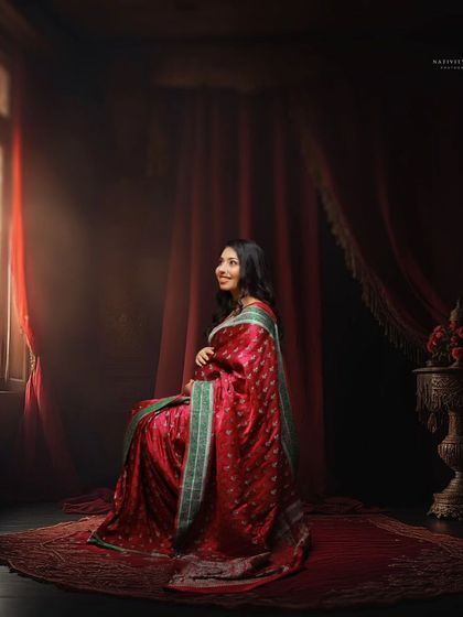 A regal portrait of a mother-to-be in a red saree, seated against a rich, dark backdrop. The side lighting from the window creates a dramatic, painterly effect.