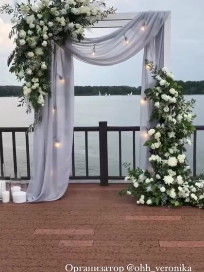 A stunning photo booth setup by the water, with a grey-draped arch and an abundance of white flowers.