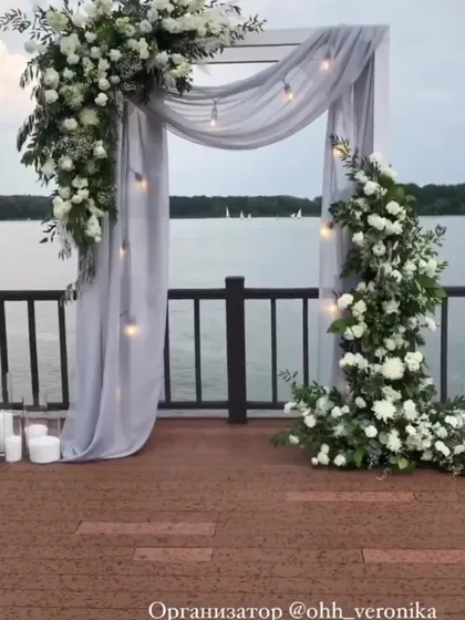 A romantic photo spot by the water, with a simple frame draped in grey fabric and adorned with white floral arrangements.