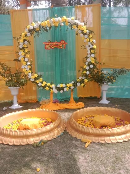An outdoor Haldi ceremony setup with two golden lotus-shaped urlis. The backdrop is a simple floral ring against light green and yellow drapes, perfect for a minimalist yet elegant event.