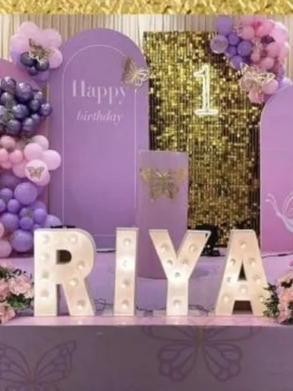 A pretty butterfly-themed first birthday for Riya. The decor features a lavender backdrop with a gold shimmer panel, marquee letters spelling out the name, and butterfly accents.