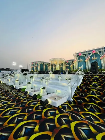 A low-angle view of the expansive royal stage, showing the detailed patterned carpet leading up to the tiered white steps and the grand teal structures. The scale of the setup is designed to be awe-inspiring.