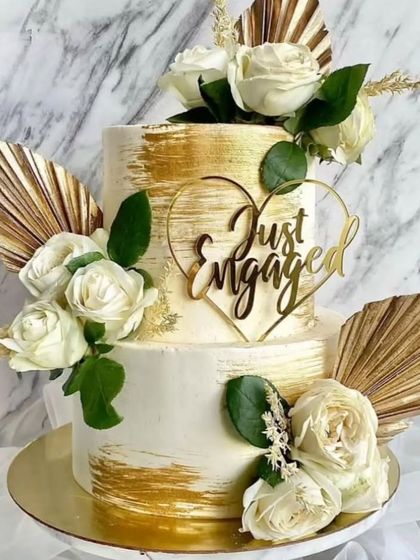 An inspiration photo of a white and gold two-tiered engagement cake with white roses and dried palm leaves.