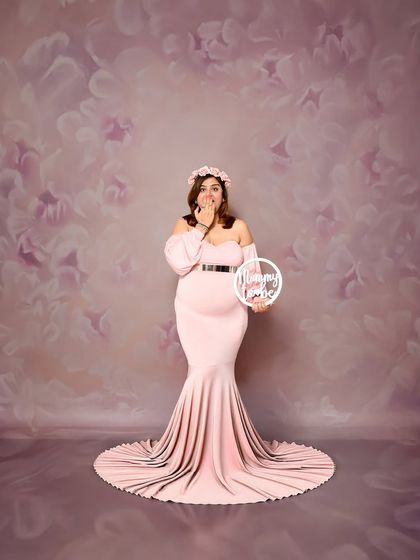 A fun and playful solo portrait of the mom-to-be holding a "Mommy to be" sign. The soft pink gown and matching floral backdrop create a sweet and charming look.
