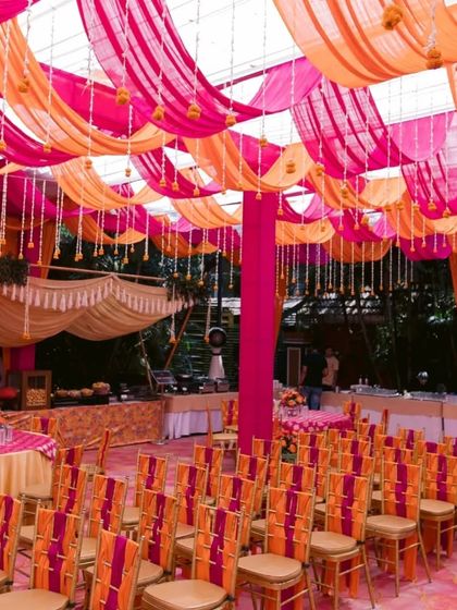 Guest seating at a colorful mehendi event. The chairs are decorated with striped orange and pink sashes, matching the vibrant draped ceiling.