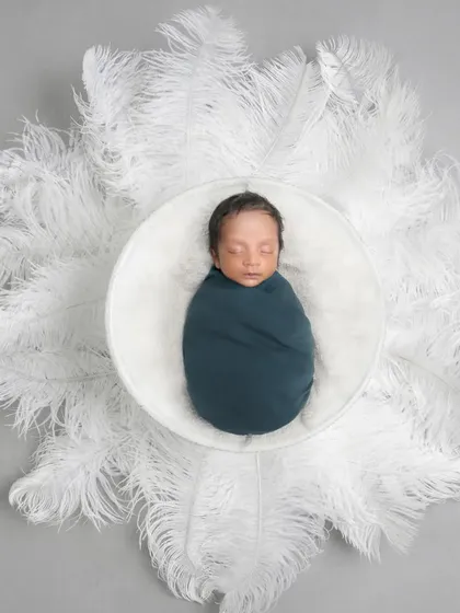 A swaddled newborn sleeping in a white bowl surrounded by a halo of large white feathers.